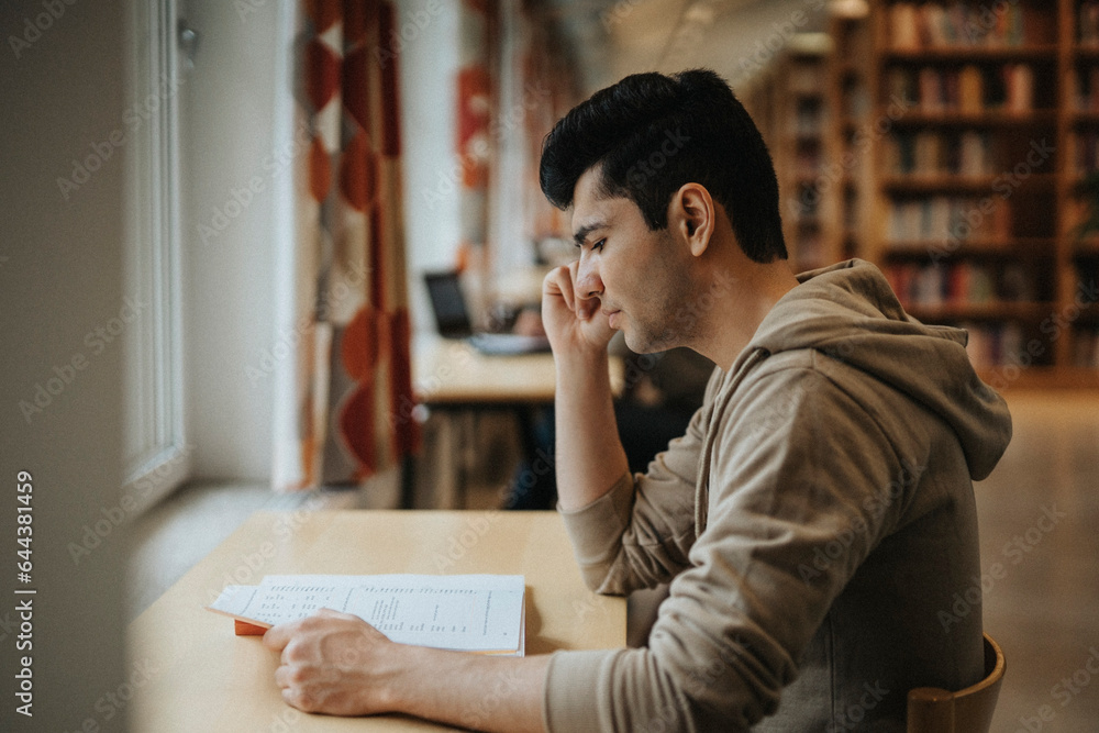 Side view of male student reading book at table in library Side view of male student reading book at table in library