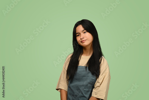Asian teenage girl wearing apron standing against on green background © Prathankarnpap