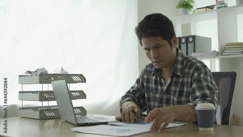 Young asian businessman working at home calculating financial graph data on laptop and checking with finance documents investment and business strategy and summarized in chart Young asian businessman working at home calculating financial graph data on laptop and checking with finance documents investment and business strategy and summarized in chart