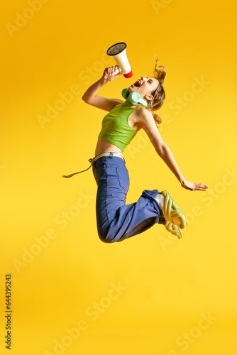 Full-length image of active, positive, young women in stylish clothes jumping with megaphone against yellow studio background. News. Concept of human emotions, youth, fashion, lifestyle, ad