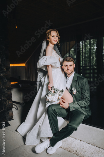 A stylish groom in a green suit and a beautiful smiling bride in a white dress are hugging while sitting on a sofa in a hotel room. Wedding photography, portrait, lifestyle.