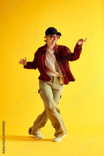 Full-length image of young woman in stylish clothes dancing and having fun against yellow studio background. Hipster. Concept of human emotions, youth, fashion, lifestyle, ad