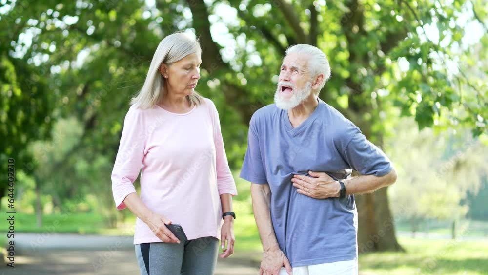 Sick senior gray haired man having a heart attack during morning training in an urban city park. Mature old wife supports her husband and calls for help online through the mobile application on phone Sick senior gray haired man having a heart attack during morning training in an urban city park. Mature old wife supports her husband and calls for help online through the mobile application on phone