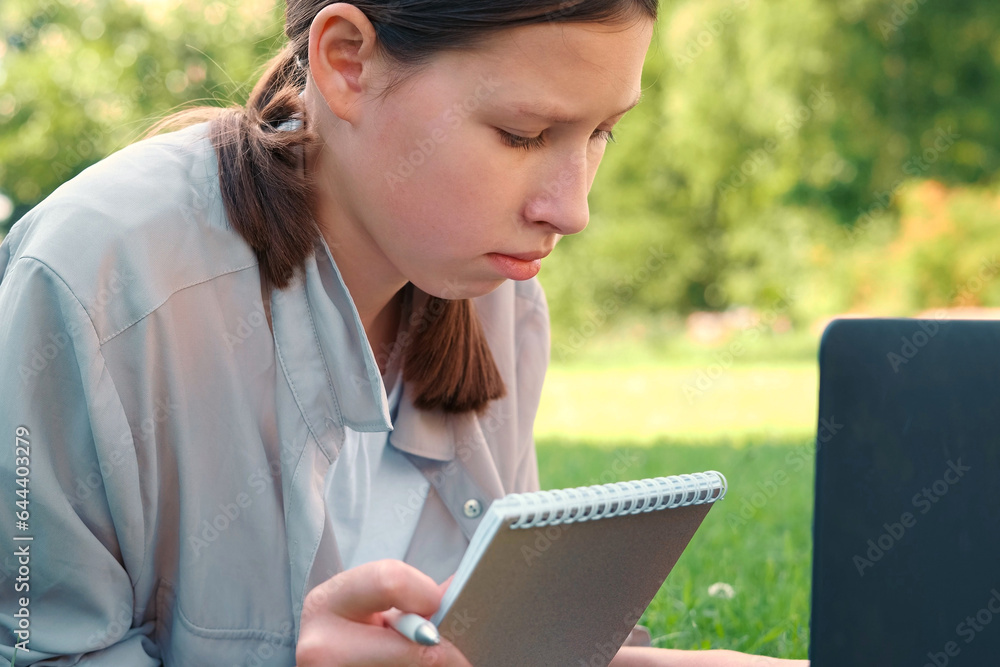 Fototapeta premium Teenage schoolgirl studying reading her books, tablet and notebook, sitting outdoors. Back to school. Student girl lying on the green grass using laptop in the college yard or park. Distance learning.