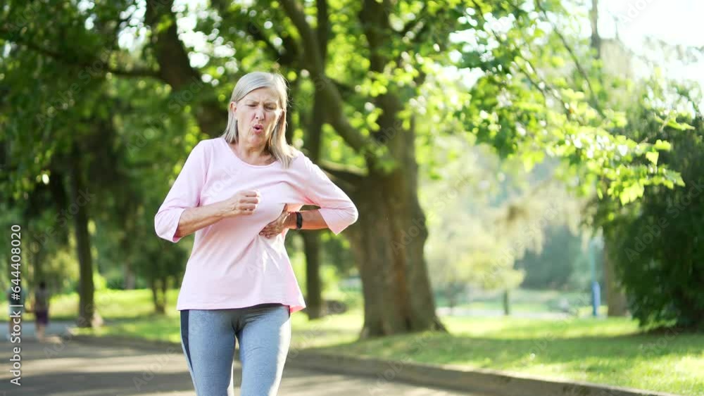 Senior adult active gray haired woman has a heart attack while running in an urban city park. Sick mature old female runner suffers from tension in the chest and holds the sore spot with her hand Senior adult active gray haired woman has a heart attack while running in an urban city park. Sick mature old female runner suffers from tension in the chest and holds the sore spot with her hand