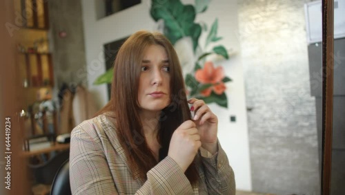 Reflection portrait in mirror of dissatisfied young woman touching hair in slow motion sitting in beauty salon. Unsatisfied sad Caucasian client indoors