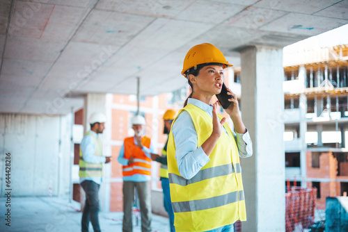 Caucasian businesswoman talk on phone while work. Gorgeous caucasian female architect in yellow west and with protective helmet on standing at construction site and using smart phone.