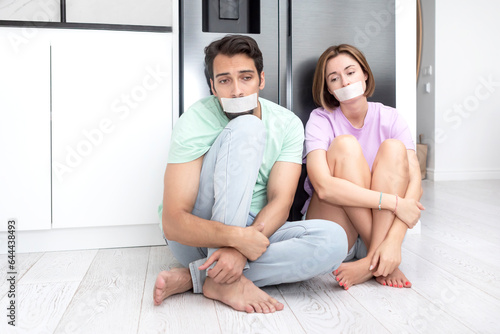Fotografija Hungry couple with taped mouths sitting close to the refrigerator and trying to