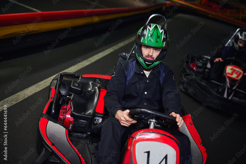 focused man in sportswear and helmet driving sport car for karting near friend on indoor circuit