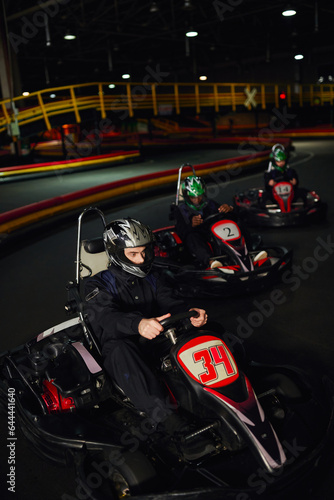 focused man driving go kart near diverse drivers in helmets on indoor circuit, adrenaline and sport