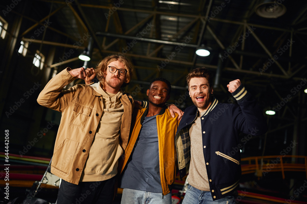 three interracial male friends smiling and standing near racing cars inside of indoor karting track