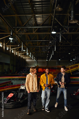 cheerful diverse friends chatting and walking near racing cars inside of karting track, motorsport