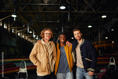 multicultural male friends smiling and standing near racing cars inside of indoor karting track