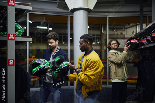 three interracial friends in casual clothes choosing helmets for karting, go-cart concept
