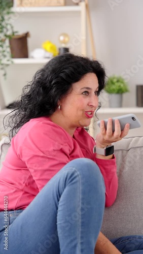 Middle aged woman sending voice message on phone while sitting on sofa