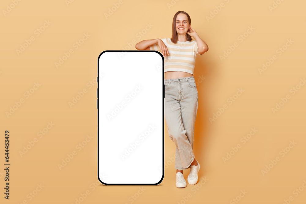 Full length portrait of smiling satisfied woman standing near big mobile cell phone with blank screen workspace area isolated on beige background space for promotional text. Full length portrait of smiling satisfied woman standing near big mobile cell phone with blank screen workspace area isolated on beige background space for promotional text.