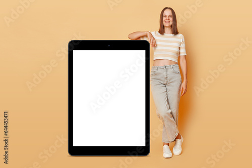 Full length portrait of smiling joyful woman standing near big digital table with white blank screen advertisement area isolated on beige background space for promotional text.