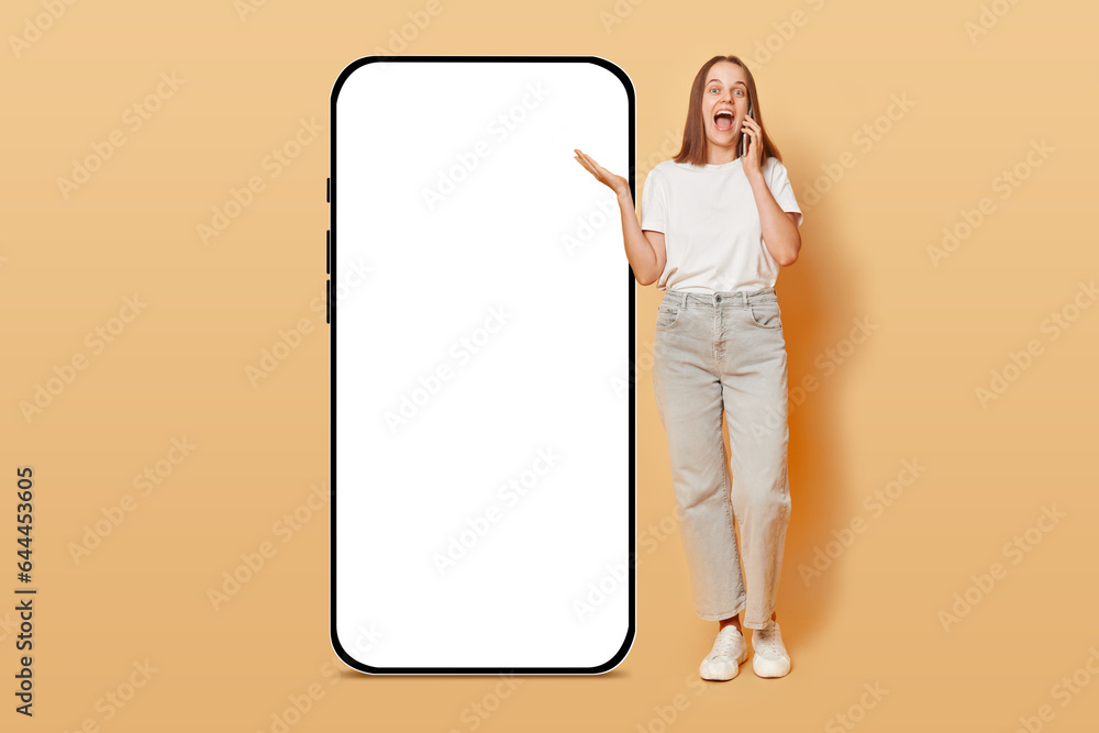 Cheerful woman talking on phone standing near big smartphone with empty screen isolated over beige background, brown haired girl communicating via cellphone, mockup. Cheerful woman talking on phone standing near big smartphone with empty screen isolated over beige background, brown haired girl communicating via cellphone, mockup.