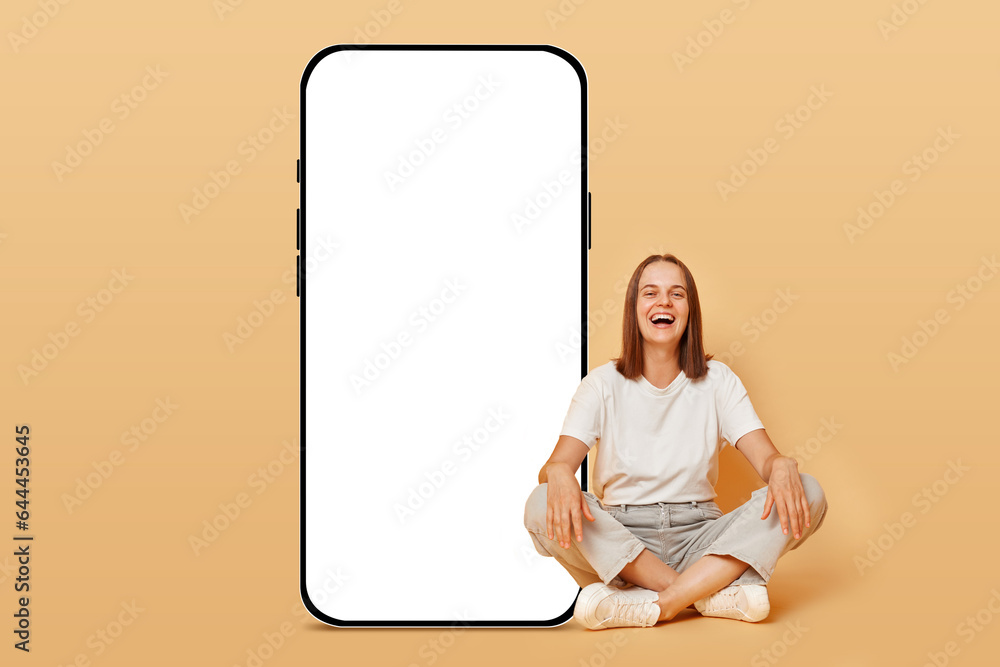 Full body of young happy smiling woman in T-shirt and jeans sitting with crossed legs near big huge blank screen smartphone mockup advertisement area isolated obeige background