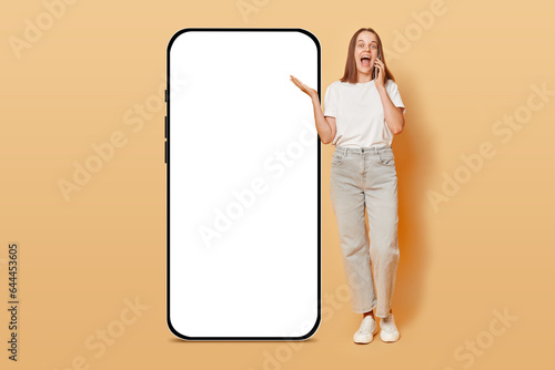 Cheerful woman talking on phone standing near big smartphone with empty screen isolated over beige background, brown haired girl communicating via cellphone, mockup.