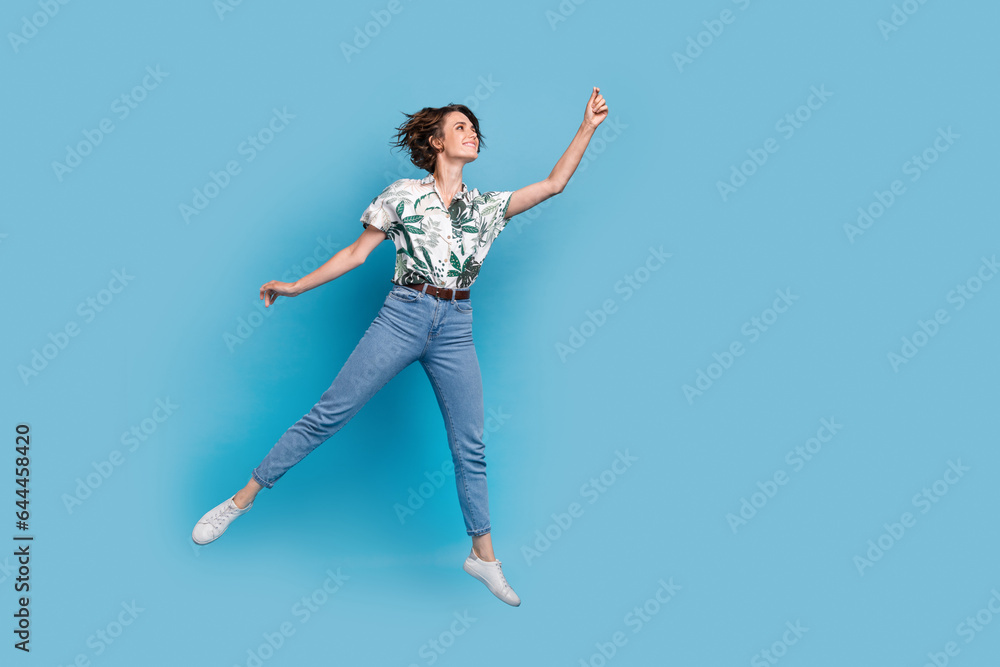 Full length photo of carefree dreamy lady wear print shirt jumping high holding empty space isolated blue color background Full length photo of carefree dreamy lady wear print shirt jumping high holding empty space isolated blue color background