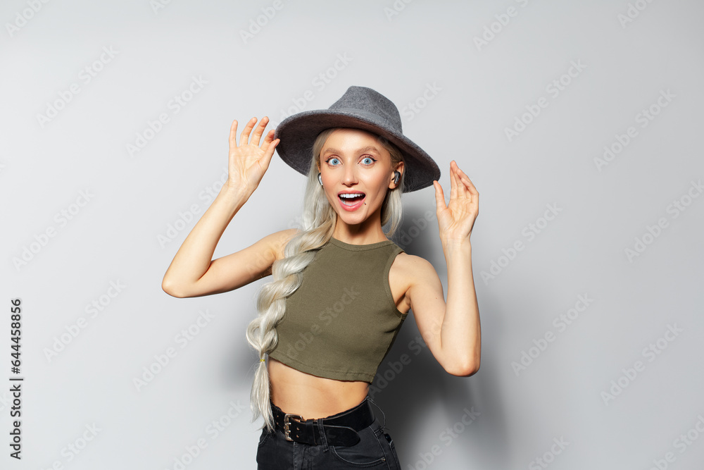 Studio portrait of attractive laughing blonde girl with wireless earbuds in ears, on white background. Wearing grey hat and green shirt. Studio portrait of attractive laughing blonde girl with wireless earbuds in ears, on white background. Wearing grey hat and green shirt.
