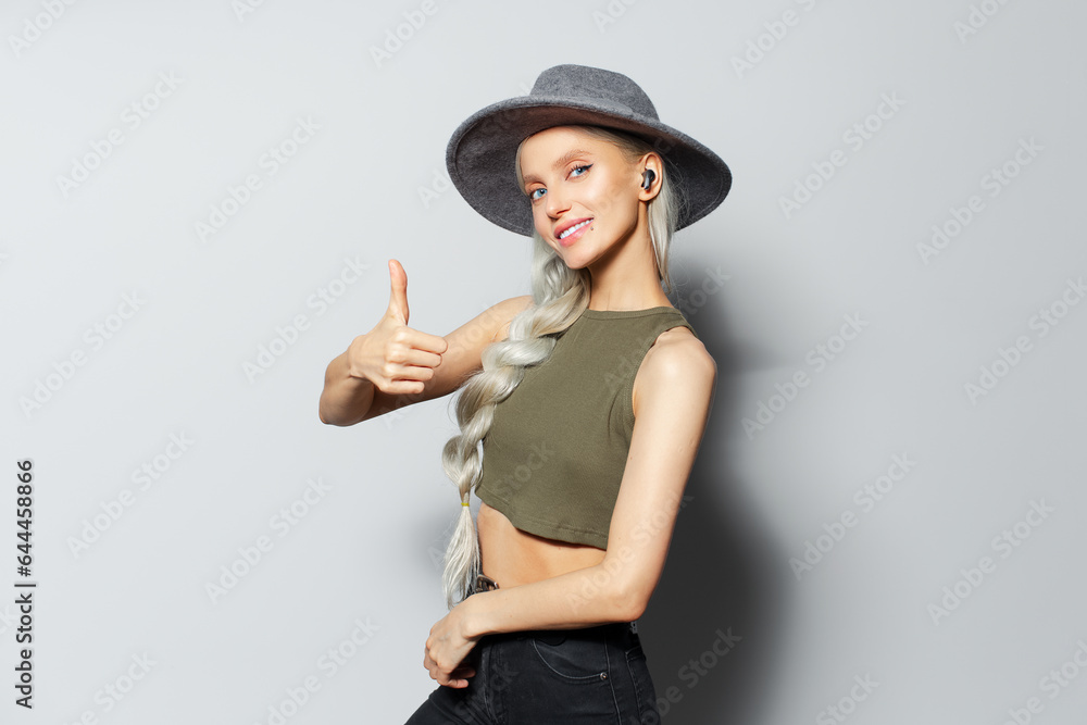 Studio portrait of attractive happy blonde girl with wireless earbuds in ears, showing thumb up, on white background. Wearing grey hat and green shirt. Studio portrait of attractive happy blonde girl with wireless earbuds in ears, showing thumb up, on white background. Wearing grey hat and green shirt.