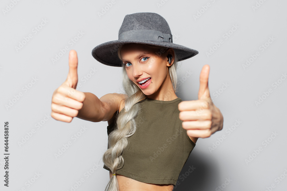 Studio portrait of attractive happy blonde girl with wireless earbuds in ears, showing thumbs up, on white background. Wearing grey hat and green shirt. Studio portrait of attractive happy blonde girl with wireless earbuds in ears, showing thumbs up, on white background. Wearing grey hat and green shirt.