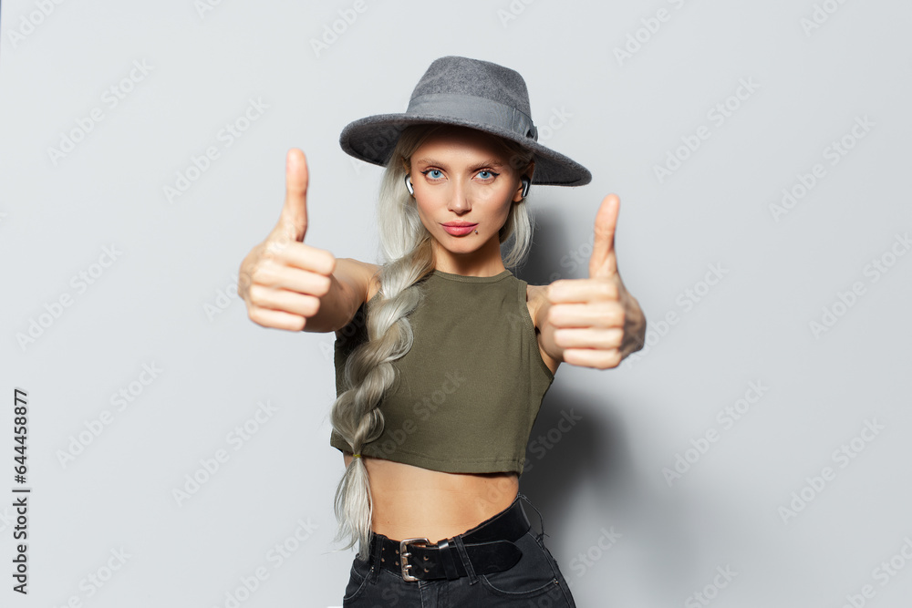 Studio portrait of attractive serious blonde girl with wireless earbuds in ears, showing thumbs up on white background. Wearing grey hat and green shirt. Studio portrait of attractive serious blonde girl with wireless earbuds in ears, showing thumbs up on white background. Wearing grey hat and green shirt.