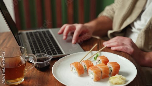 Close-up of a young handsome man dressed in a beige shirt scrolling the social media feed on a laptop, eating sushi with salmon photo