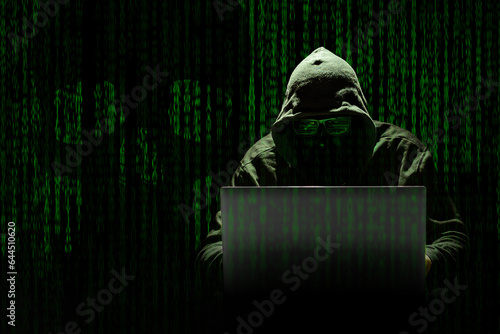 Slika na platnu Un hacker encapuchado trabaja en un ciberataque sobre un fondo de código informático donde se aprecia una calavera