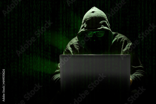 Leinwand Poster Un hacker encapuchado cometiendo un ciberataque con un portátil, sobre un fondo de código informático estilo Matrix