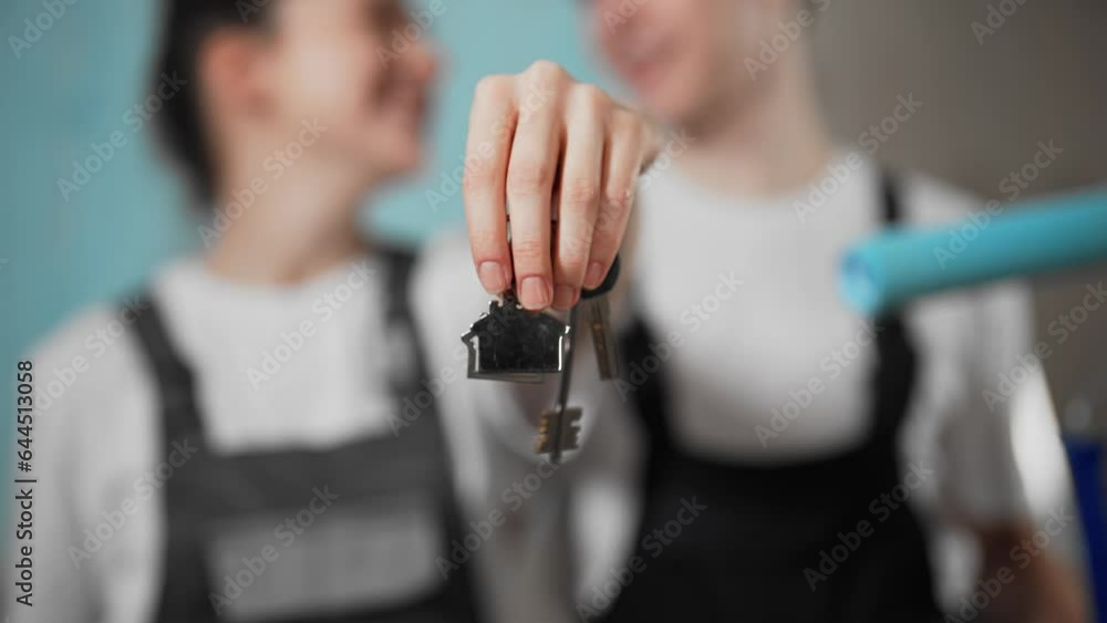 Couple shows keys keychain from new apartment. Happy family in working clothes makes renovation in house flat, paints walls in blue color uses paint roller. Purchase, real estate, mortgage concept. Couple shows keys keychain from new apartment. Happy family in working clothes makes renovation in house flat, paints walls in blue color uses paint roller. Purchase, real estate, mortgage concept.