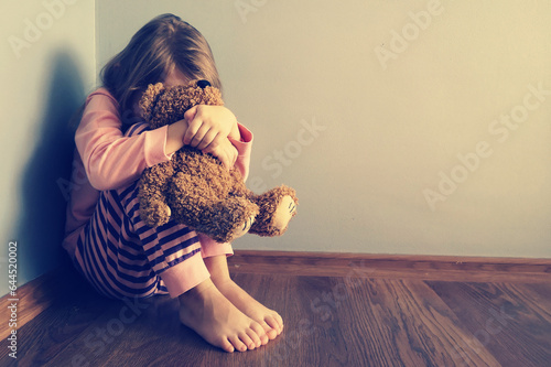 The child is sitting sad scared on the floor in the corner and hugs a toy. Little girl crying with her head down. Concept: No Domestic Violence and Child Abuse. Mistakes of parents in raising children