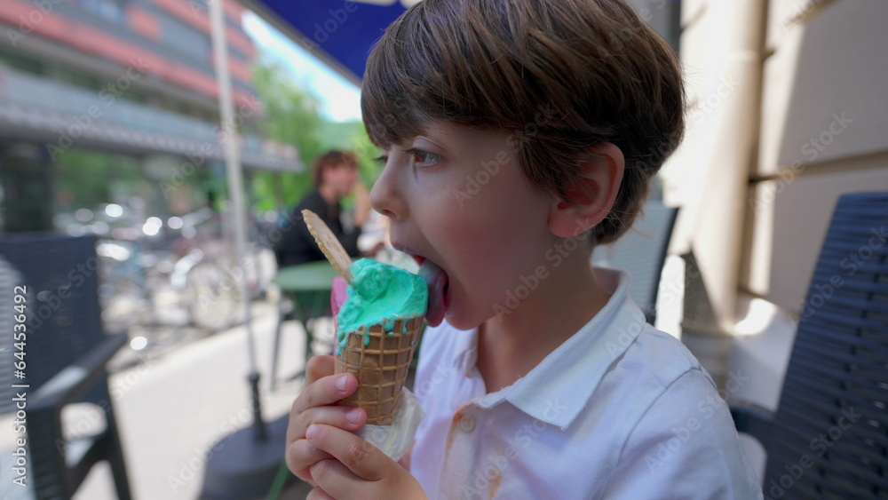 Small boy eating colorful icecream cone outside at parlor shop's sidewalk. Profile face of kid licking sweet dessert indulgence