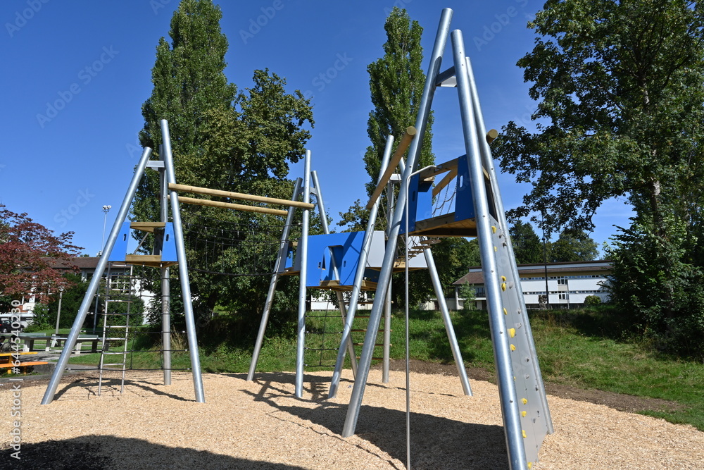 Playground for children with various equipment made of metal, wood and ropes. On the ground there is mulch because of safety reasons. There are trees on the background. 