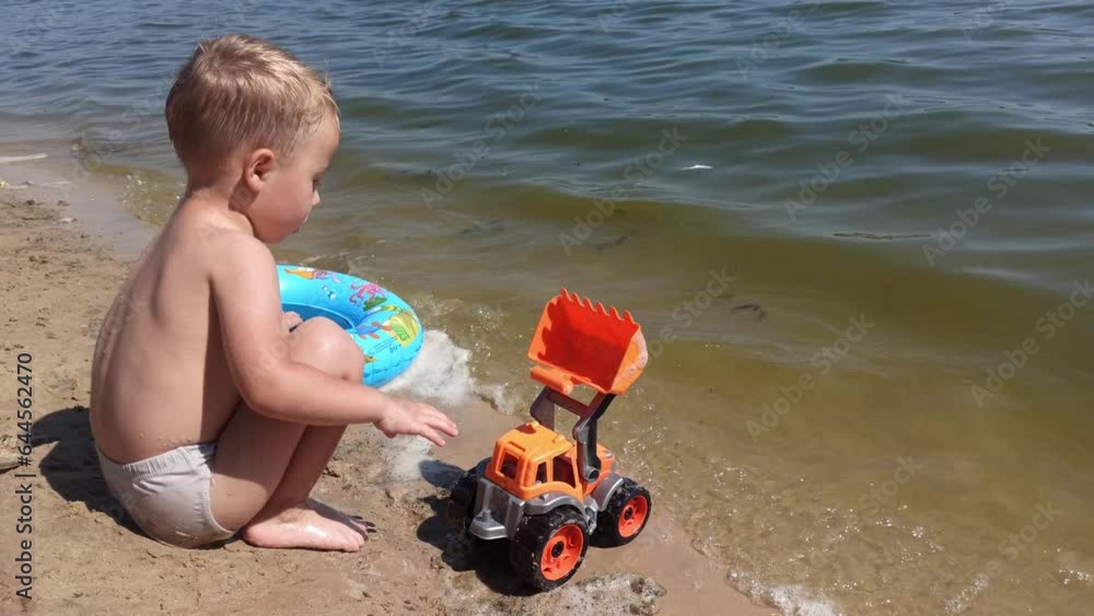 Kid boy at the river coast plays with sand and toy. Summer leisure time. Family trip to the seaside during holidays. 4k footage Kid boy at the river coast plays with sand and toy. Summer leisure time. Family trip to the seaside during holidays. 4k footage