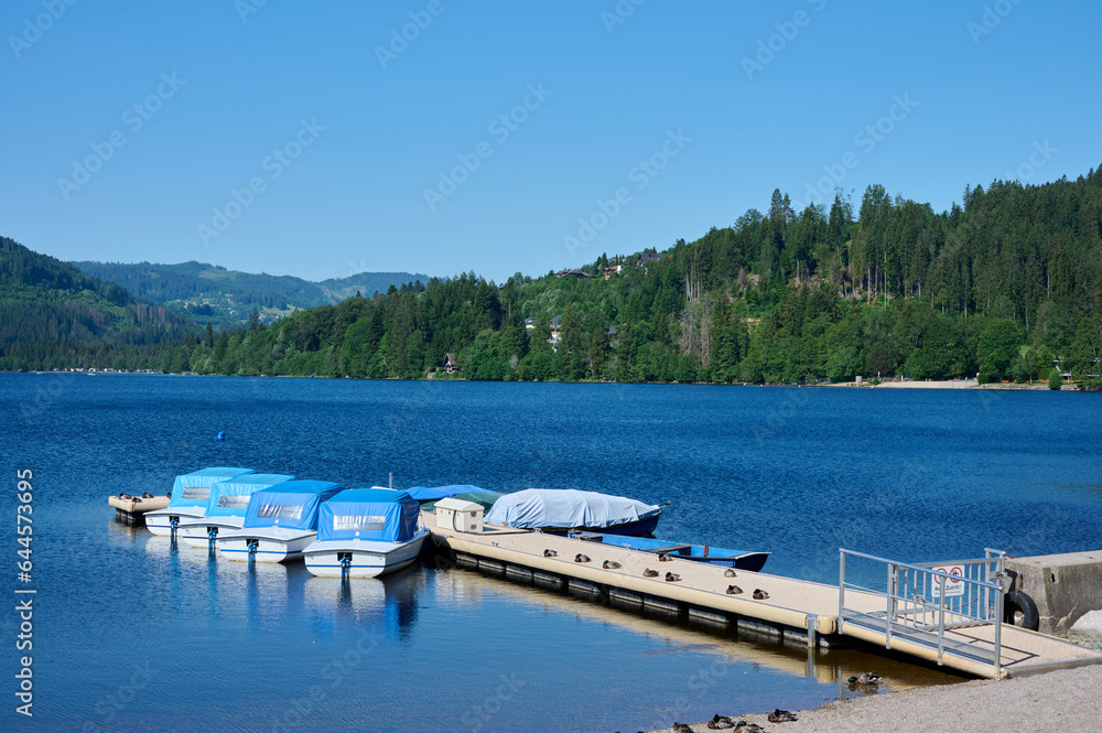 Fototapeta premium Titisee with blue water and boats on the jetty, blue sky, mountains and green trees