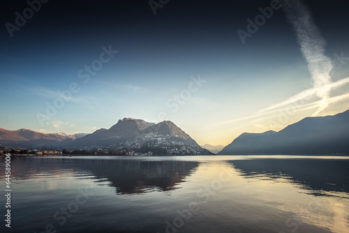 Fototapeta Naklejka Na Ścianę i Meble -  Sunrise panorama over Lugano lake with mountain and clouds reflections. Swiss and Italian mountains in the background some of which are illuminated by the sun. Lugano, Ticino, Switzerland