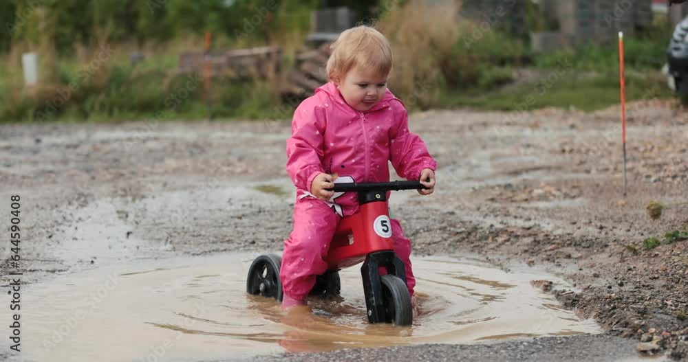 Small oneyear old baby girl and mother playing in mud puddles after heavy rain. Jumping, throwing rocks and examine new . Wearing pink purple raincoat and Wellingtons. Riding a red bike, dogs around Small oneyear old baby girl and mother playing in mud puddles after heavy rain. Jumping, throwing rocks and examine new . Wearing pink purple raincoat and Wellingtons. Riding a red bike, dogs around
