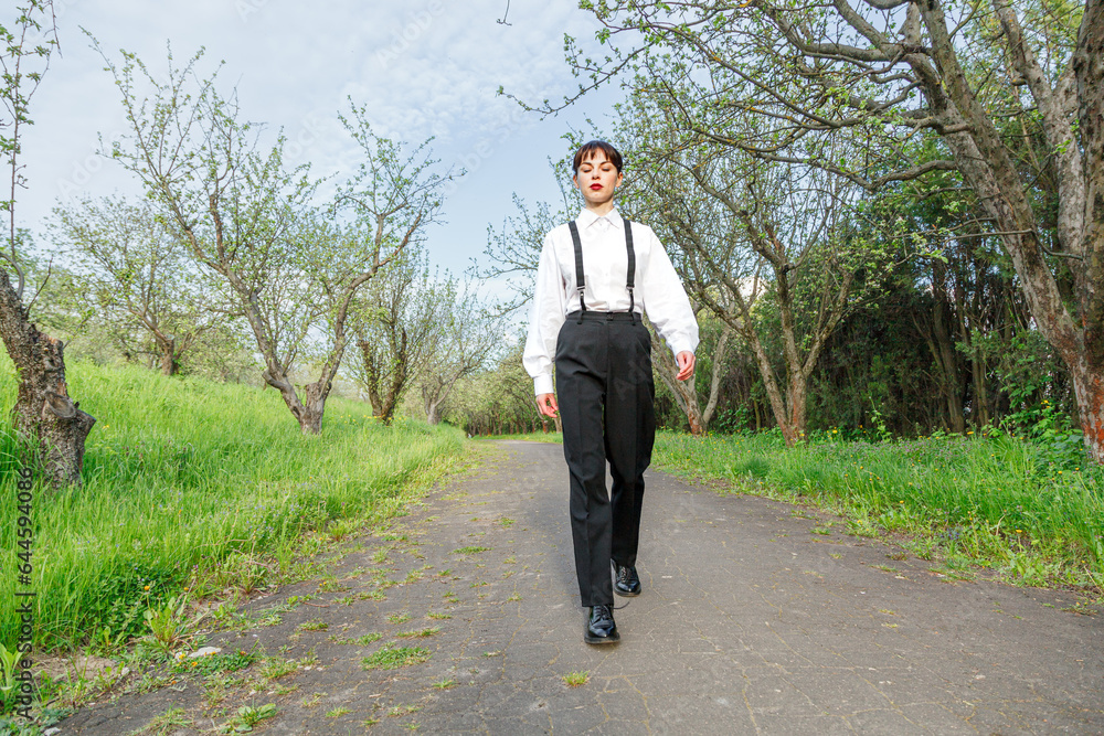 A beautiful girl in a white shirt, in black trousers with suspenders against the background of the sky and green grass