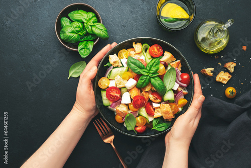 Hands holding plate with  panzanella salad with stale ciabatta bread, colorful tomatoes, soft cheese, red onion, olive oil, sea salt and green basil, black table background, top view photo