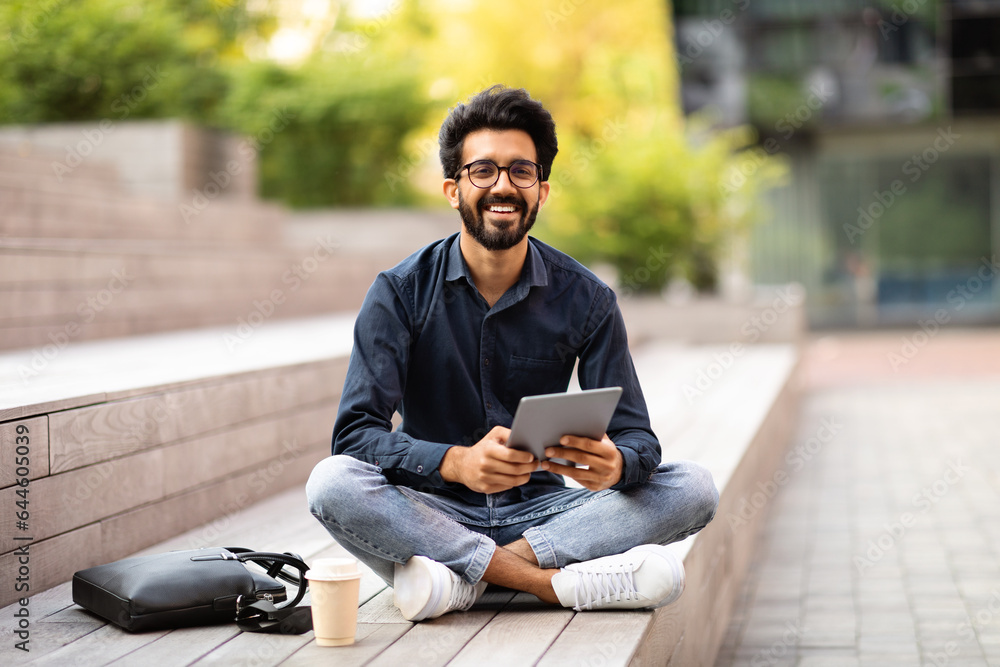 Obraz premium Indian guy sitting outdoor with digital tablet, having coffee break