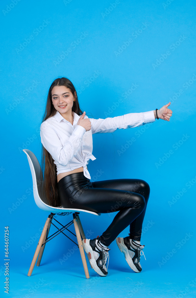 Side view of joyful beautiful woman showing thumb up with outstretched hand, while sitting in modern chair and smiling at camera, isolated on blue studio background. Concept of gesturing. Side view of joyful beautiful woman showing thumb up with outstretched hand, while sitting in modern chair and smiling at camera, isolated on blue studio background. Concept of gesturing.