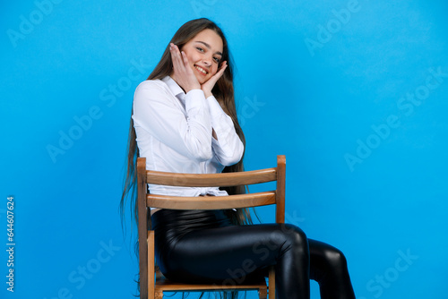 Beautiful young woman in white shirt, holding hands on face, while smiling at camera. Portrait of happy female model sitting on chair and posing, isolated on blue studio background. Concept of posing. photo