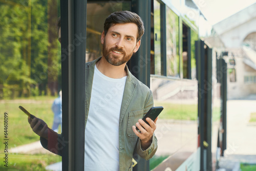 Smiling handsome businessman with cellphone getting off public transport. Front view of busy bearded male passenger with mobile phone looking away, while leaving city bus. Transport, business concept. photo