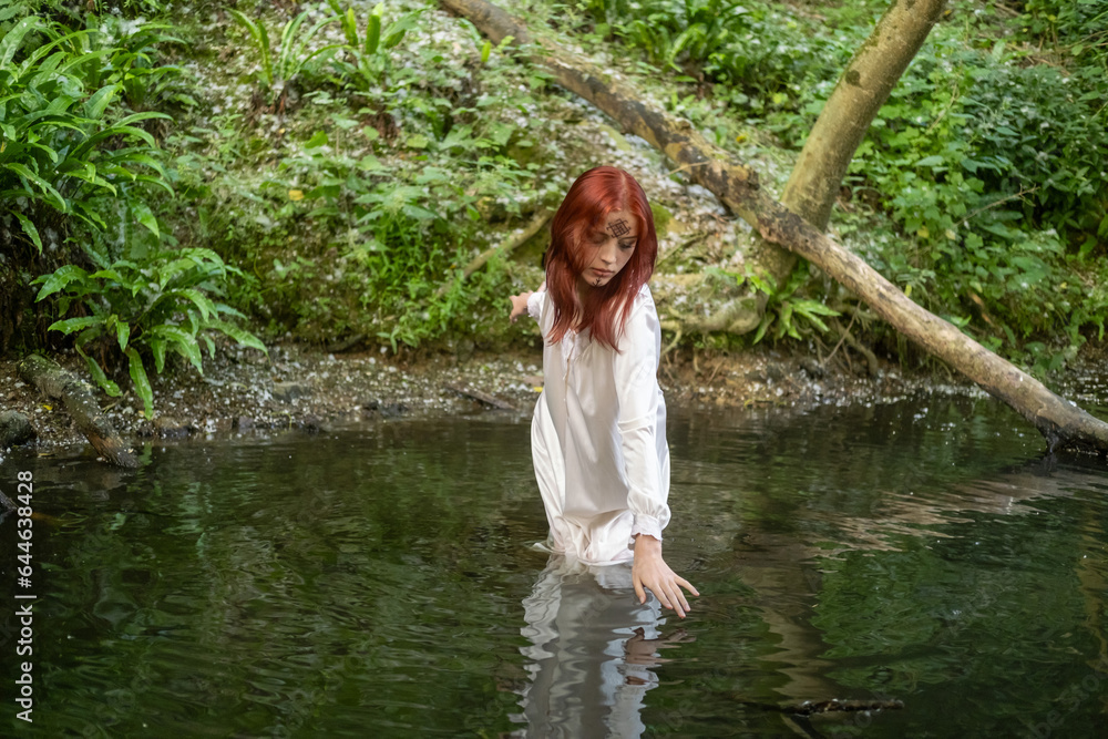 Fototapeta premium A young teenage girl in a white dress stands in a lake against a background of green foliage. day Ivan bathed.