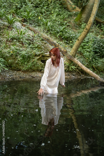A young teenage girl in a white dress stands in the water, in a lake against a background of green foliage. day Ivan bathed.