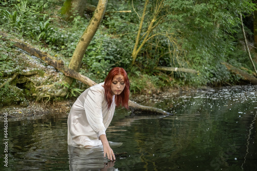 A young teenage girl in a white dress stands in the water, in a lake against a background of green foliage. day Ivan bathed.