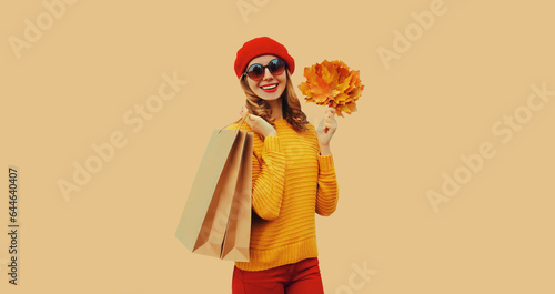 Autumn color style outfit, portrait of beautiful young woman with shopping bag holds yellow maple leaves wearing red french beret hat, knitted sweater on beige studio background